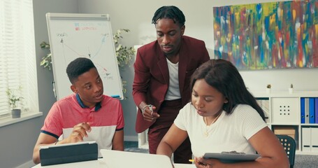 Elegantly dressed in a maroon suit, a dark-skinned manager approaches a woman approving his documents, signing, working as a team, co-workers helping themselves