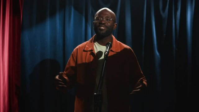 Medium low key shot of male Black stand-up comedian in glasses, bright orange shirt and white t-shirt actively gesturing, telling jokes and laughing while performing on small stage