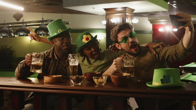 Medium lowkey shot of cheerful Caucasian man in glasses taking selfie with African American friends while hanging out with beer at bar counter in evening on St Patricks day