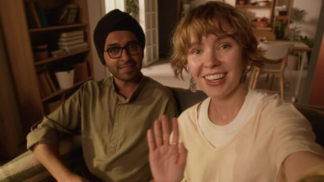 POV UGC shot of young happy blonde Caucasian lady waving and talking to camera, her Indian boyfriend in turban and glasses sitting behind her on sofa at home and smiling, both looking at camera