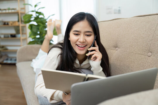 Happy young woman lying on a couch, talking on the phone and reviewing notes, expressing joy and engagement. - Powered by Adobe