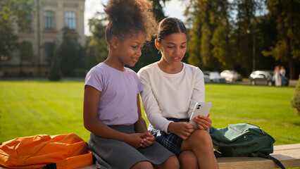 Fototapeta premium Multiethnic multiracial kids little girls schoolgirls female pupils students African American friends classmates school education online study outdoors gadget addict children mobile phone social media
