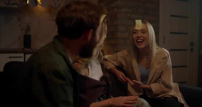 Happy blonde girl trying to guess the word on a sticker and chatting with her friends during a fun and word game at a party in the evening