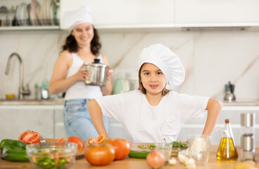 Happy little girl posing by kitchen-table while her mother standing behind in the kitchen