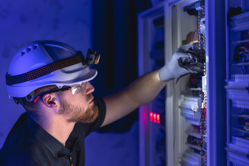 A male electrician works in a switchboard in overalls against the backdrop of emergency lighting.