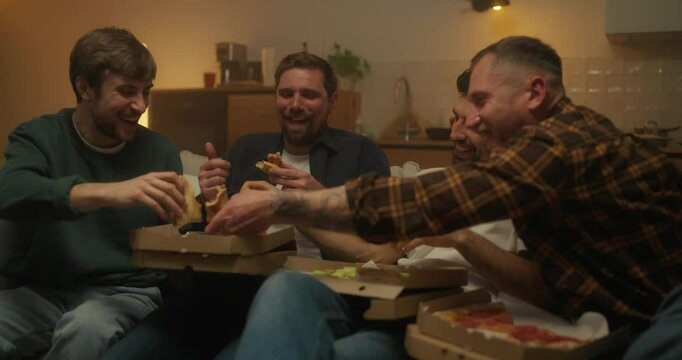 Four friends are sitting on the sofa and actively eating pizza from paper boxes while relaxing and having a bachelor party at home in the evening
