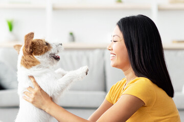 Side view of happy young chinese woman in casual outfit sitting on floor in cozy living room,...
