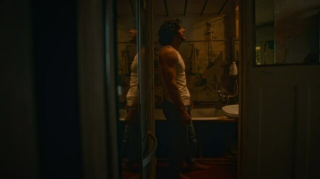 Full side shot of despondent and weakened man in undershirt and jeans looking at himself in mirror in worn out bathroom in evening