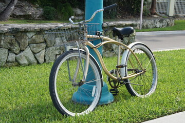 Beach cruiser bike, leaning against a turquoise pole on a patch of green grass. Classic design,...