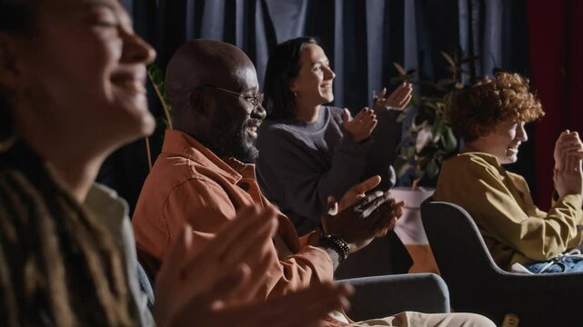 Side low key shot of Black male spectator and group of audience cheering, clapping and laughing vigorously during standup night performance