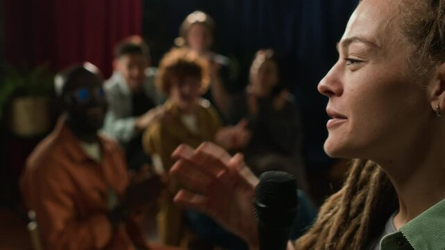 Close up side shot of Caucasian female stand-up comedian actively gesturing and joking on stage, receiving lot of applause and ovation from audience, performing in club