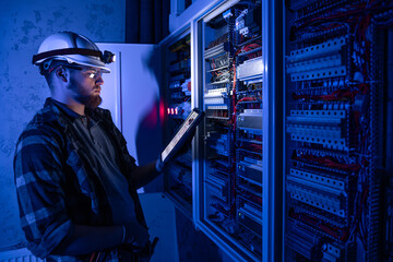 Electrician in overalls, focused on work in switchboard with fuses, using tablet.