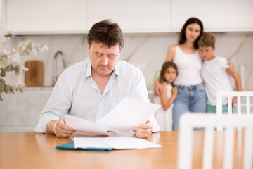 Obraz premium Worried adult man examining documents while sitting at table in kitchen with family