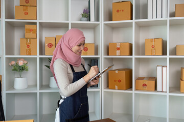 Muslim woman taking notes while managing her online business inventory at home.