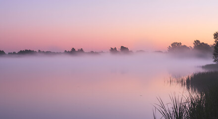 Obraz premium Soft pastel hues of a sunrise over a calm lake, with mist hovering above the water.
