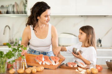 Mother with little daughter in kitchen while cooking salmon dish. Woman and girl communicate, mother explains culinary tricks and subtleties to her daughter.