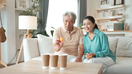 Happy elderly couple enjoying a ball toss game together at home, practicing coordination and cognitive skills for healthy aging. Grandfather and grandmother throwing ping pong in the bowl. Myrmidon.