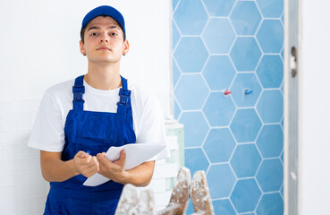 Confident young builder in blue overalls planning process of indoor building overhaul, making...