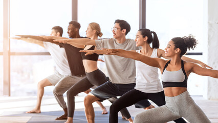 Obraz premium Multiethnic group of sporty people practicing yoga, standing in Warrior two pose in modern loft studio, side view