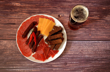 A glass of semi-dark beer and a plate with an assortment of meat snacks, croutons and cheese on a wooden table.