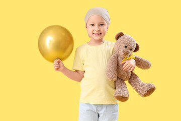 Cute little girl after chemotherapy with yellow ribbon, teddy bear and air balloon on color...