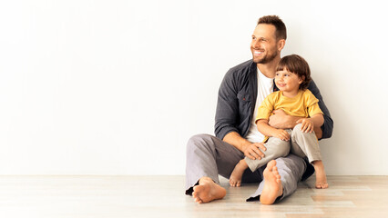 Great family ad. Happy caucasian father carrying his little son, sitting together on floor and looking aside at empty space, white wall background, mockup for text