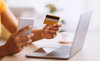 Making Financial Transaction. Closeup of unrecognizable black lady holding credit card and smartphone, sitting at desk with laptop computer, blurred background. Online Shopping And Payment Concept