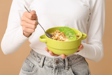 Young woman holding bowl with tasty granola and kiwi on beige background, closeup