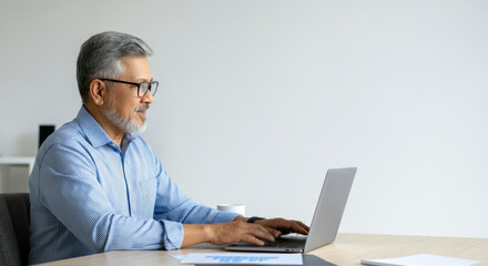 A man in a blue shirt is typing on a laptop in front of a white wall