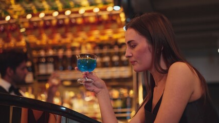 Sitting beautiful woman guest feeling bored waiting friends or couple with drinking mixed vodka in blue hawaii cocktail at luxurious counter bar for Friday meeting on bartender background. Vinosity.