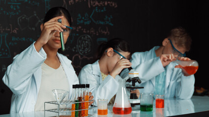 Cute girl looking under microscope while student doing experiment at blackboard with theory...