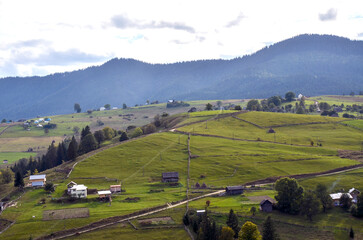 Idyllic rural scenery featuring green rolling hills spotted with small houses and farm structures, surrounded by lush greenery and distant forested mountains under a cloudy sky. Carpathians, Ukraine