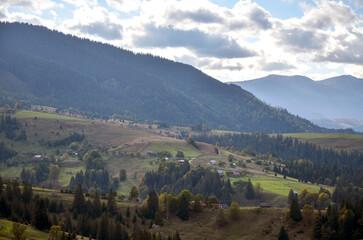 Fototapeta premium Idyllic rural scenery featuring green rolling hills spotted with small houses and farm structures, surrounded by lush greenery and distant forested mountains under a cloudy sky. Carpathians, Ukraine