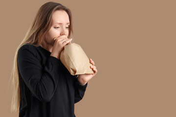 Young woman with paper bag having panic attack on beige background