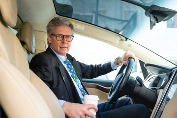 A senior businessman with grey hair and glasses sits confidently in a sleek, modern electric car,...