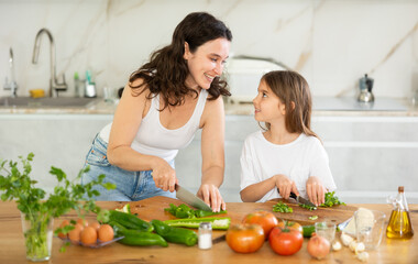 Happy mother and her little daughter are cutting fresh vegetables for salad in the kitchen