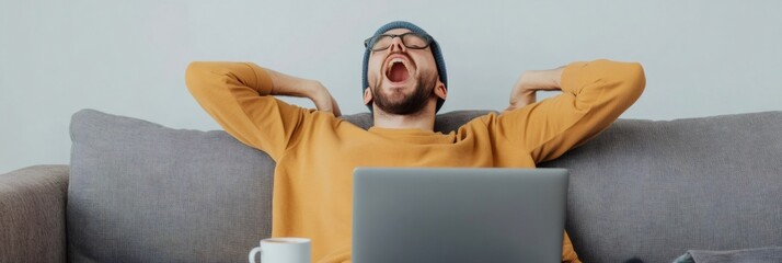 Tired man wearing glasses and a beanie yawns while sitting on a couch with a laptop, indicating stress and fatigue from multitasking and managing deadlines in a remote work environment
