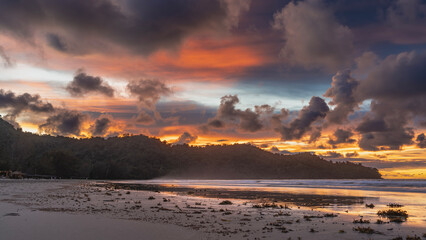A bright sunset on a tropical island. Purple clouds in the blue sky are highlighted in scarlet and orange. Bunches of algae on the beach. Reflection on wet sand. A hill in the distance. Malaysia.