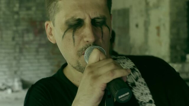 Passionate singer sings rock music into the mic, full of energy. Behind him, drummer pounds intense beats. Clad in punk fashion, they perform inside an abandoned structure, bringing the place to life