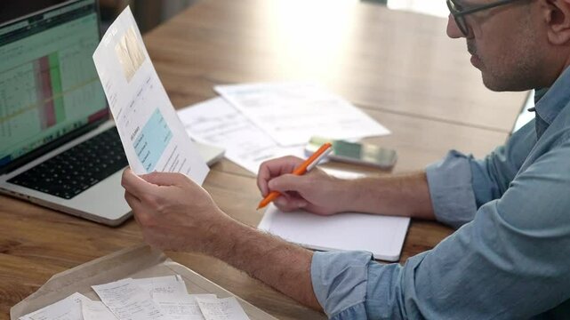 Middle-aged man reviews utility bill taking notes in paper notebook, male office worker calculates expenses related to office services preparing financial reports for company