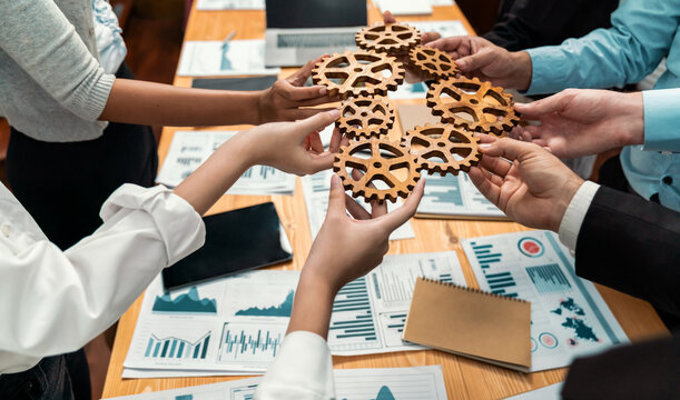 Group of multiracial business people joining gear wheels together over table as effective unity and productive teamwork concept. Efficient system business team process for solution solving. Meticulous