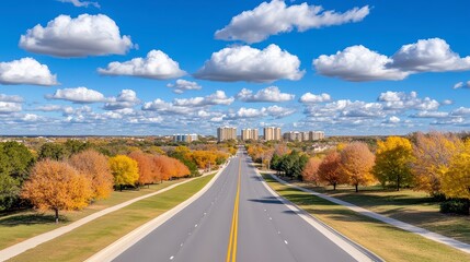 Obraz premium Tranquil Autumnal Road Leading to Cityscape Under Blue Sky with Fluffy Clouds