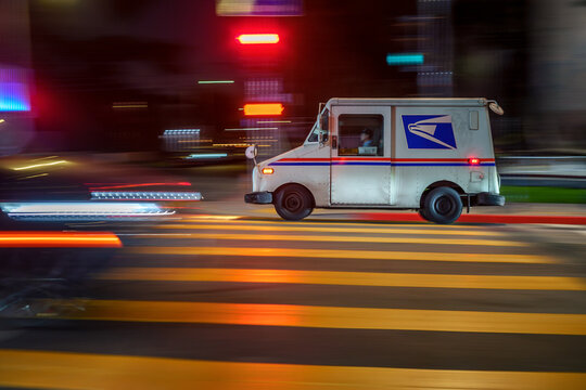 LOS ANGELES, CA - February 19, 2025: USPS delivery truck rushing through city traffic, crossing illuminated street intersection at night.