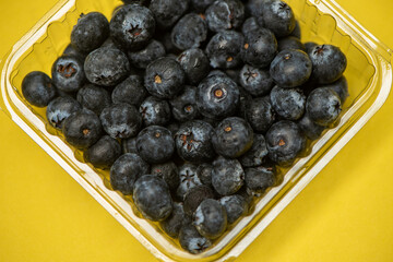 Fresh blueberries on a yellow surface.Blueberries have a dark blue color and a slightly rough texture.  A clear plastic container filled with blueberries.