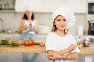 Girl poses against background of kitchen interior. Small scullion with ladle stands in kitchen after cooking