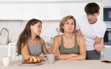 Sad mom sits silently in kitchen, adult children, guy and girl sympathize and comfort woman....