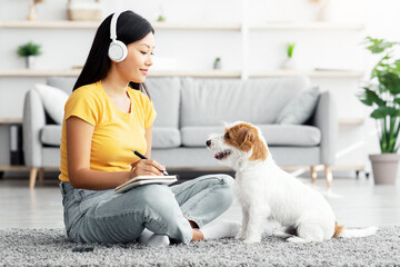 Happy dog fluffy jack russel terrier sitting by cheerful female owner young asian woman in casual sitting on floor in living room, using wireless headset and taking notes, learning foreign language