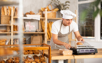 Interested focused young seller in striped apron and chef hat carefully weighing fresh pastries to package for sale in rustic artisan bakery