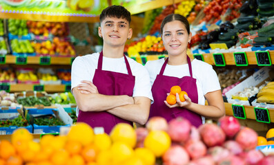 Positive happy smiling saleswoman and salesman in apron offering fresh fruits on fruit store