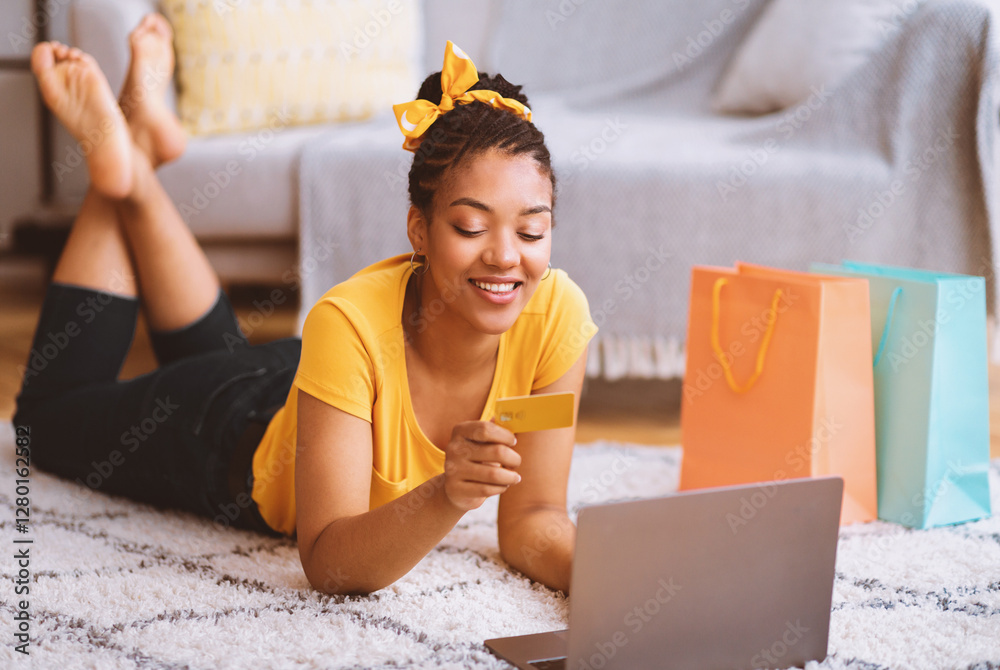 Poster Online Transaction Concept. Smiling black lady using laptop and credit card, ordering purchases via internet, lying on the floor carpet, checking debit card number and typing cvv security code